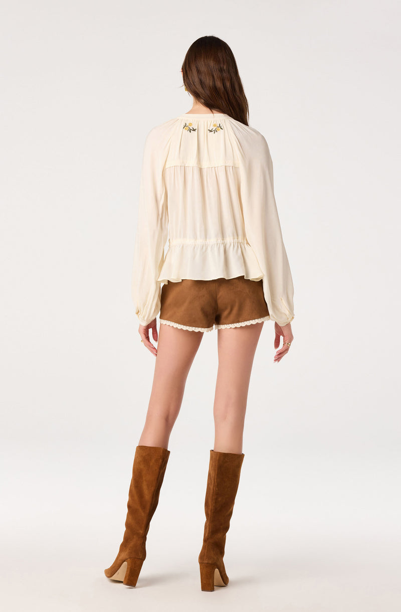 Cream blouse with embroidered details and ruffled hem, paired with brown shorts with lace trim.