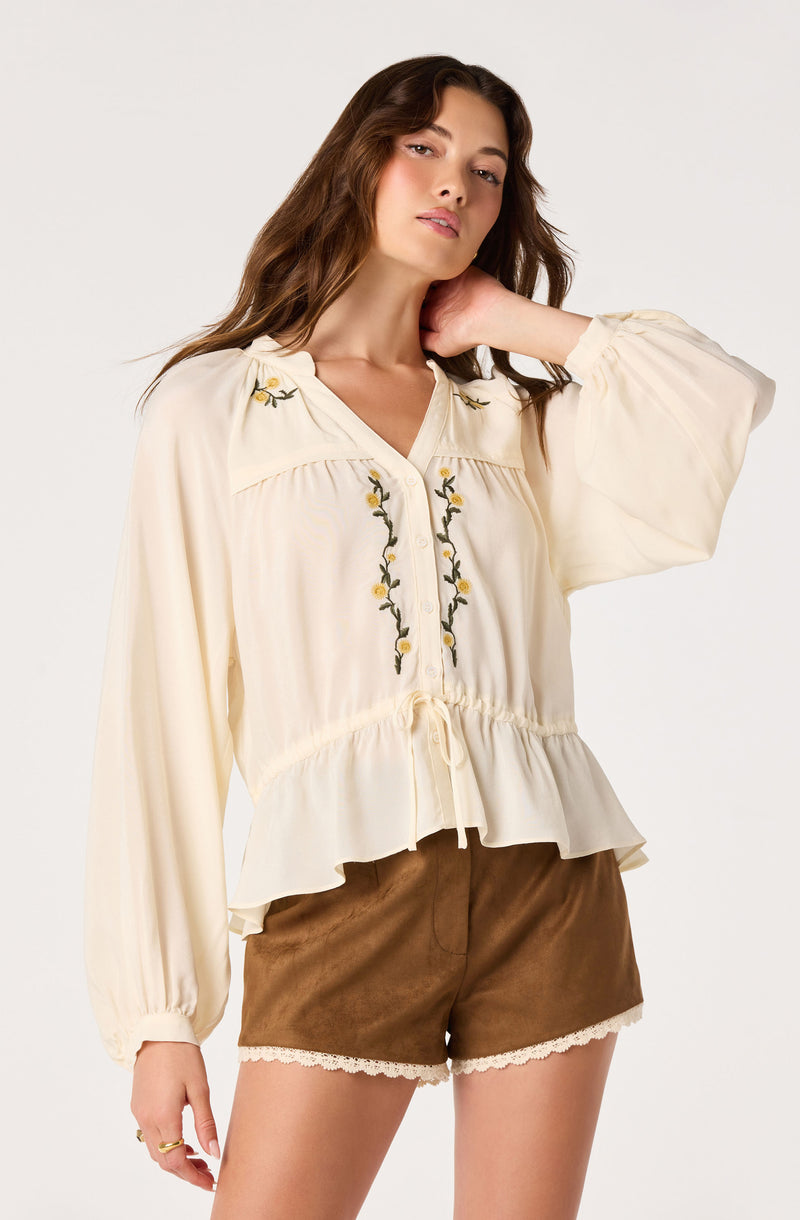 Woman in cream blouse with floral embroidery and brown shorts with lace trim.