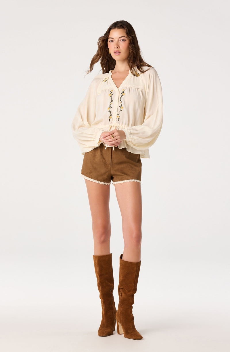 Woman wearing a cream peasant blouse with embroidered details, brown shorts, and tall brown boots.