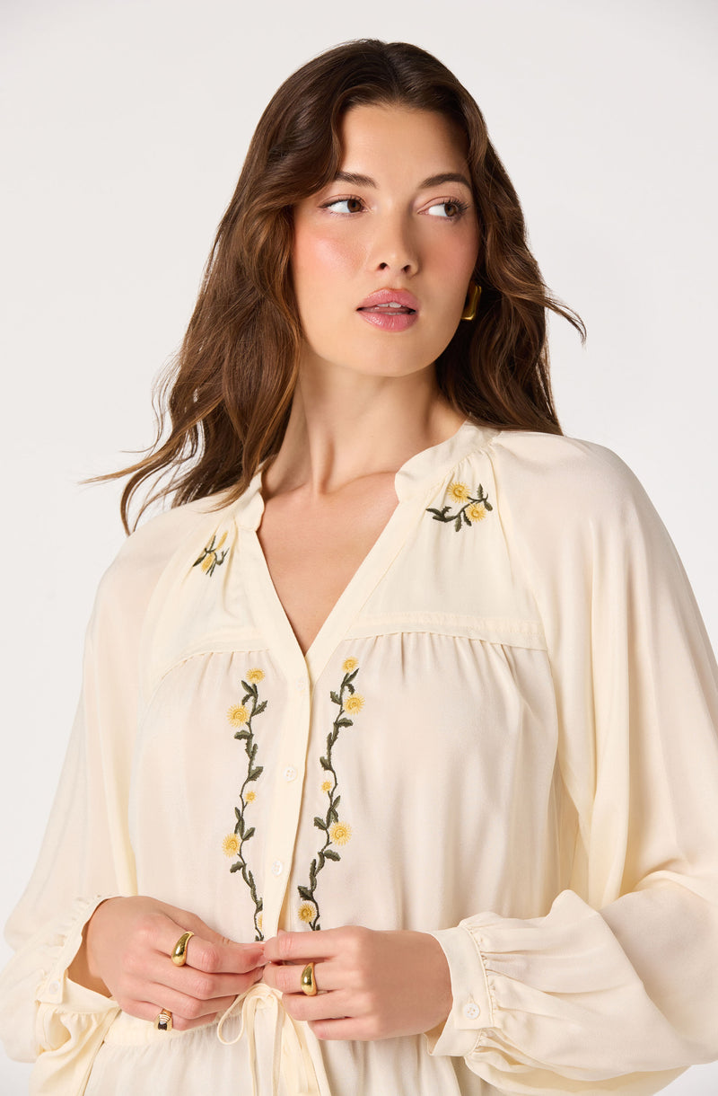 Cream-colored blouse with floral embroidery, long sleeves, and a tie at the waist.