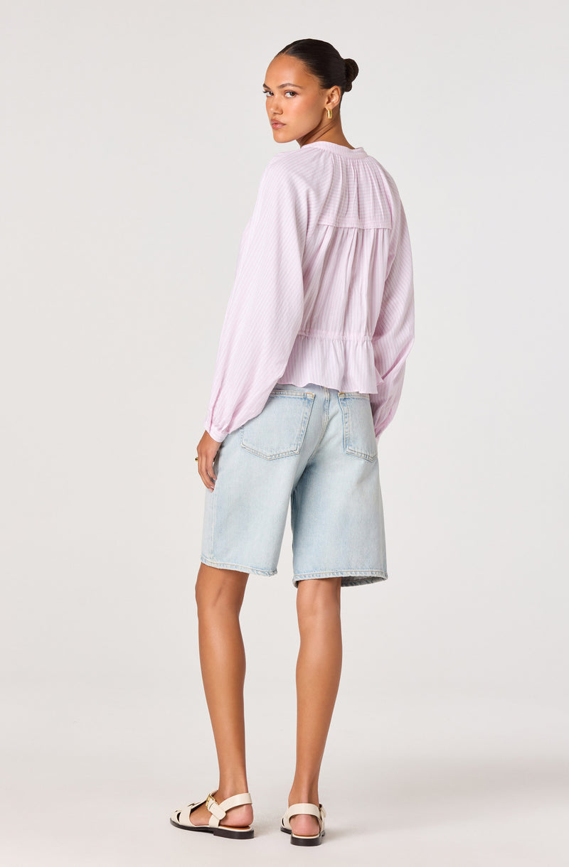 Woman wearing a light pink, long-sleeved, collared blouse with ruffle hem, and light blue denim shorts.