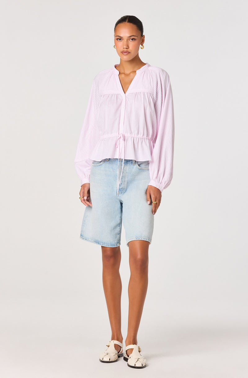 Woman wearing a pink, long-sleeved, loose blouse with a drawstring waist, light denim shorts, and white sandals.