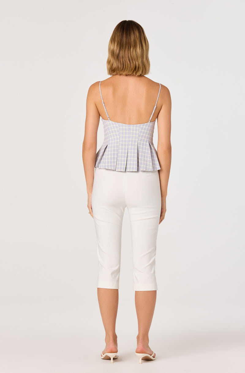 Woman wearing a light purple and yellow checkered sleeveless top with thin shoulder straps and a pleated hem, paired with white capri pants.