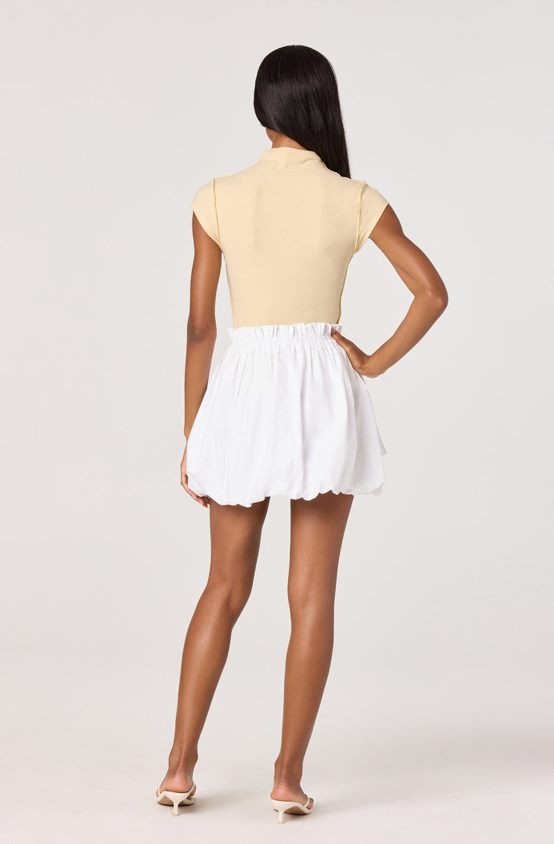 Woman in a beige sleeveless top and white elastic-waist skirt, standing with one hand on her hip.