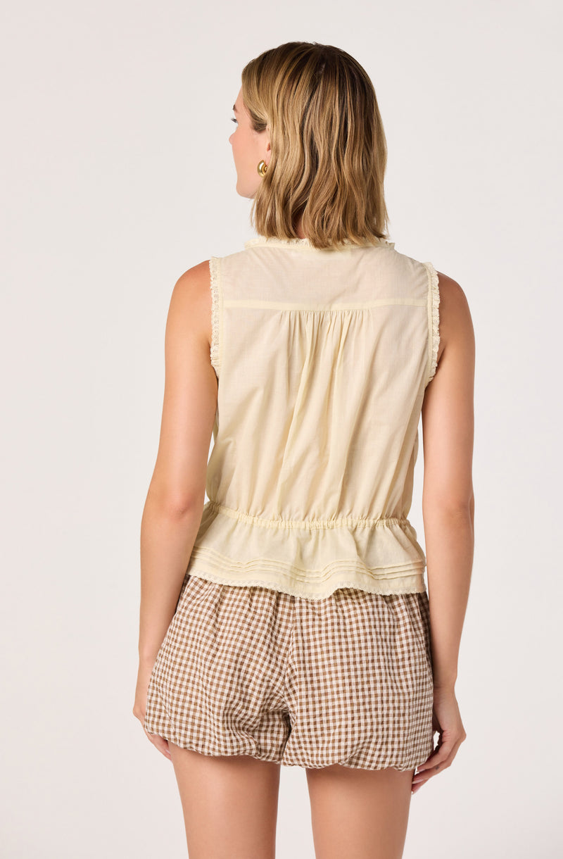 Woman wearing a sleeveless beige top with fringe details and brown checkered shorts.