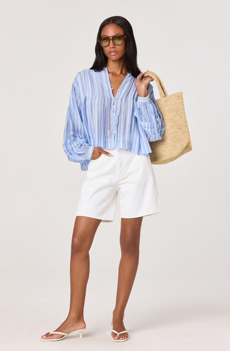 Woman wearing a blue and white striped blouse, white shorts, beige heeled sandals, and sunglasses, carrying a straw tote bag.