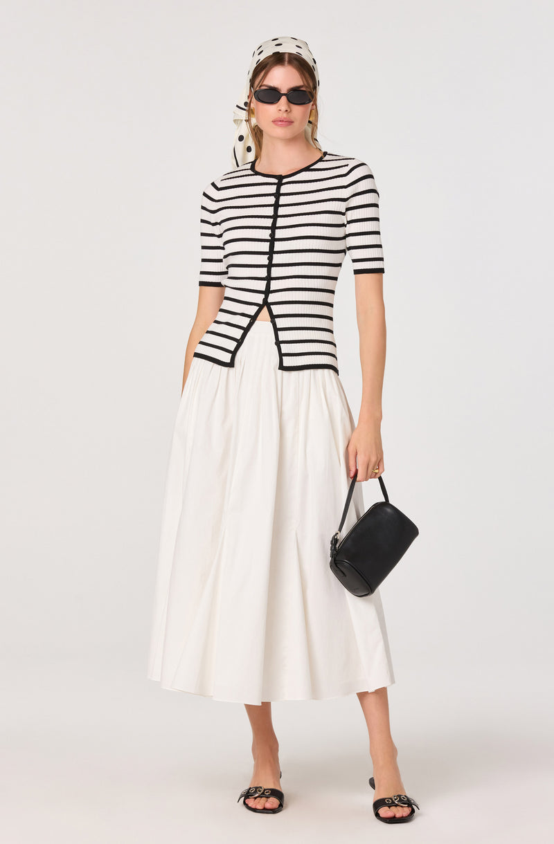 Model wearing black and white striped short-sleeve top, white wide-leg skirt, black sunglasses, polka dot headscarf, black handbag, and black sandals.