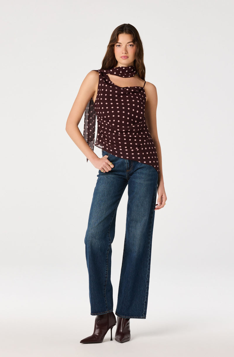 Woman wearing a sleeveless maroon top with pink polka dots and a matching scarf, blue jeans, and dark brown high-heeled boots.