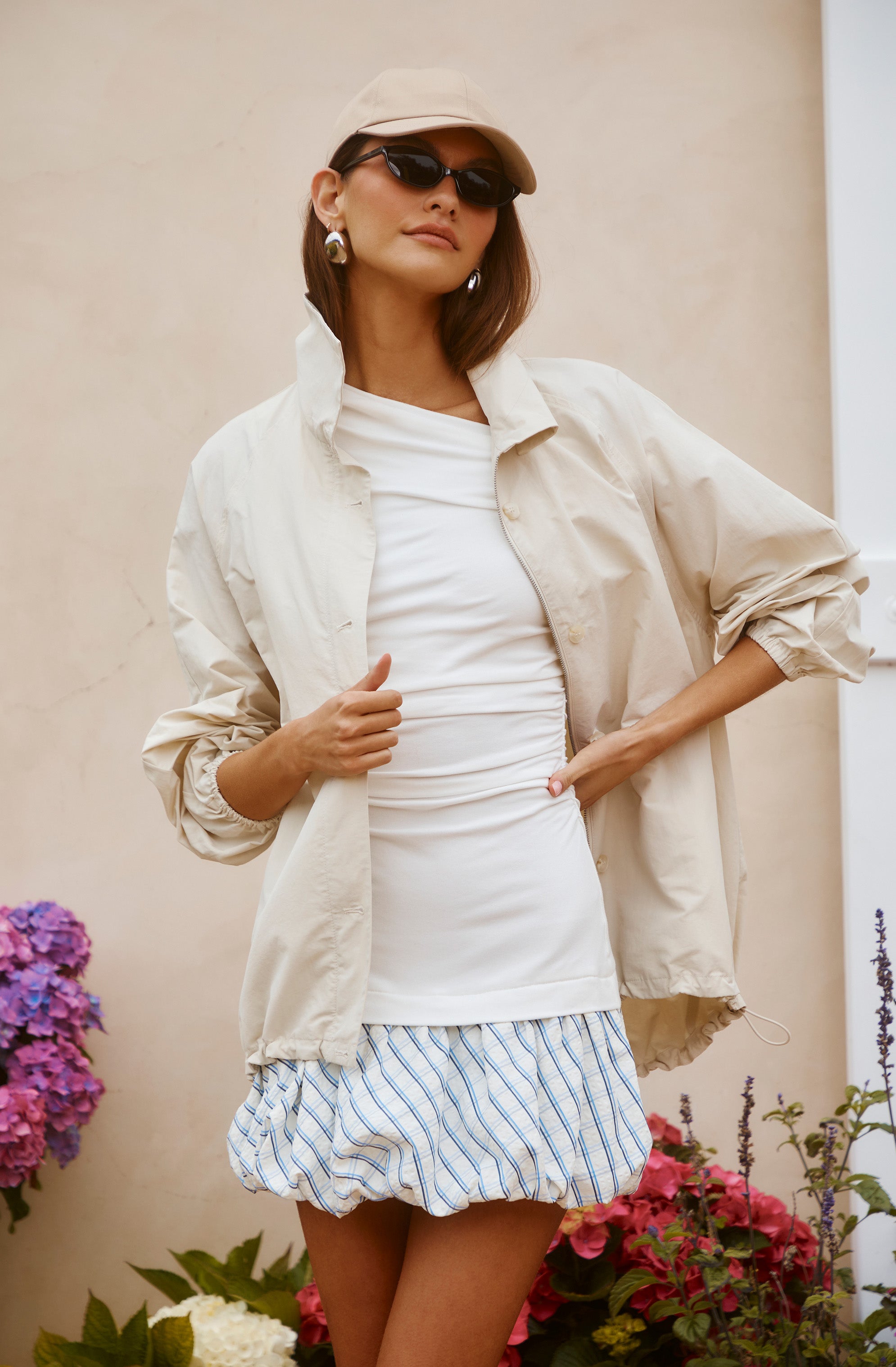 Woman wearing black sunglasses, a beige cap, beige jacket, white top, and blue striped skirt.