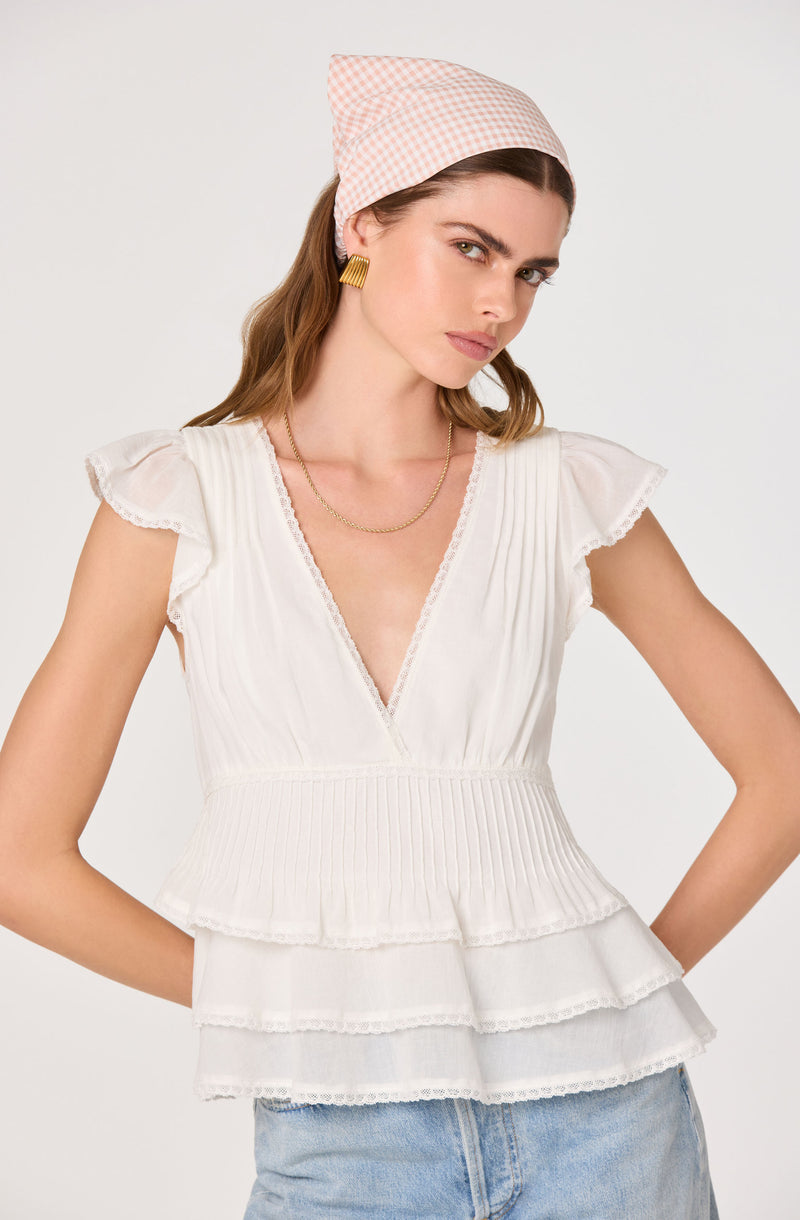 Woman wearing a white ruffled, tiered top with pleated details and a pink gingham headscarf.