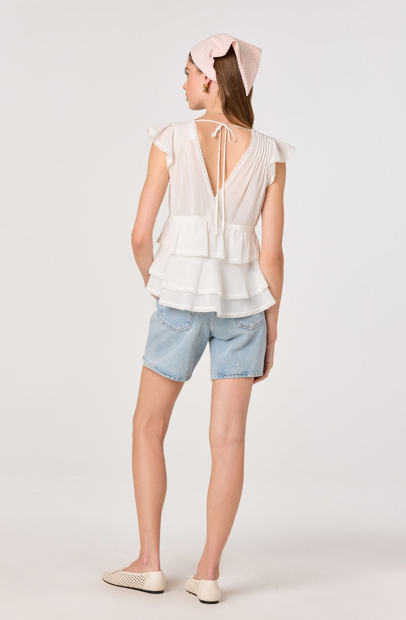 Woman wearing a white ruffled top with tiered layers, light blue denim shorts, and white flat shoes with cutout details. She has a pink and white checkered headscarf.