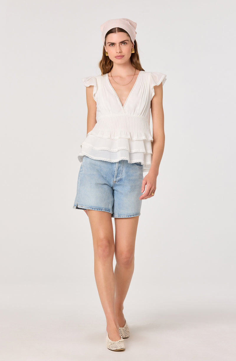 Woman wears a white layered sleeveless top, light blue denim shorts, beige shoes and a pink cap.