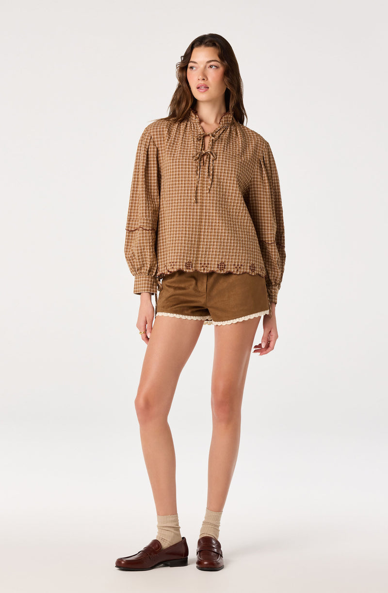 Woman wearing a brown checked blouse with a bow tie and puffed sleeves, paired with brown shorts with white lace trim.