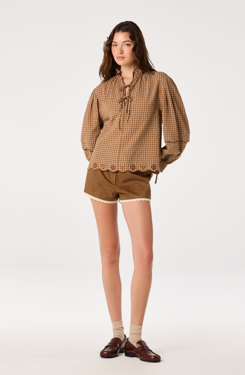 Girl wearing brown checkered blouse with puffed sleeves and bow tie, brown shorts with lace trim.
