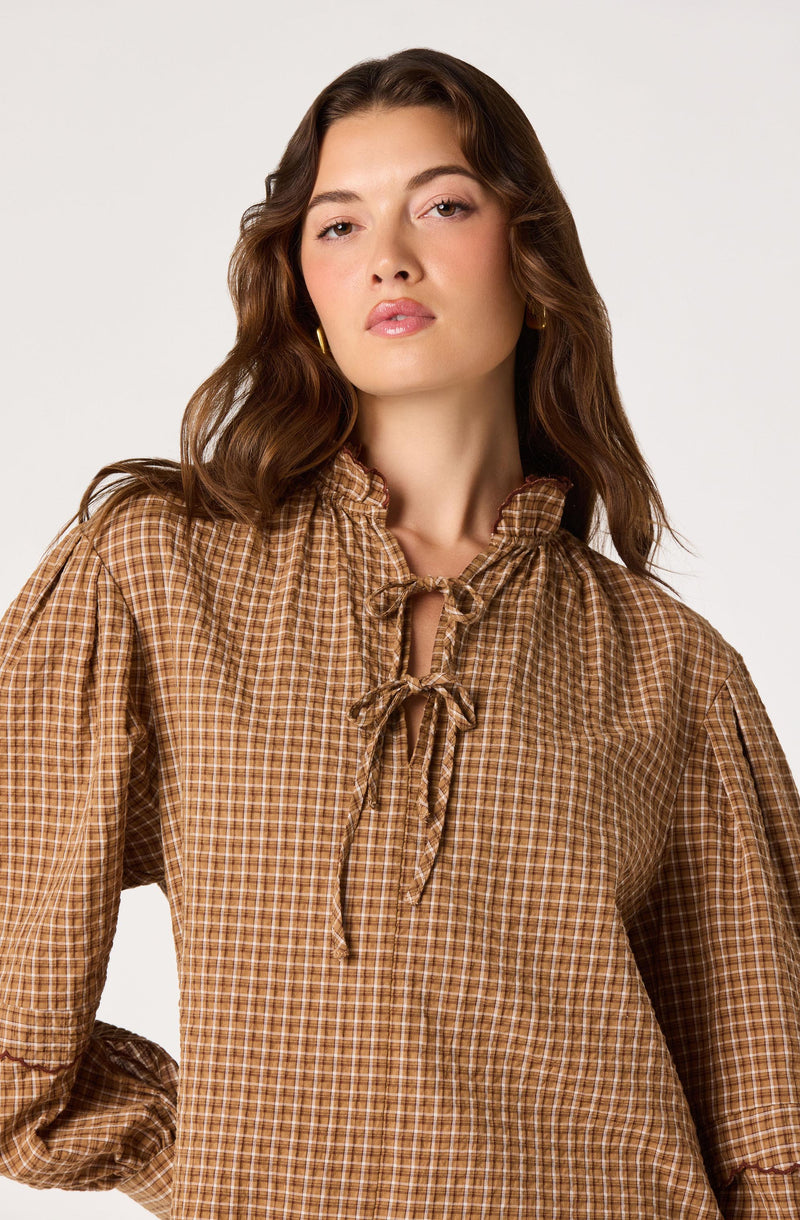 Woman wearing a brown checkered blouse with a ruffled collar and a bow tie at the neck.