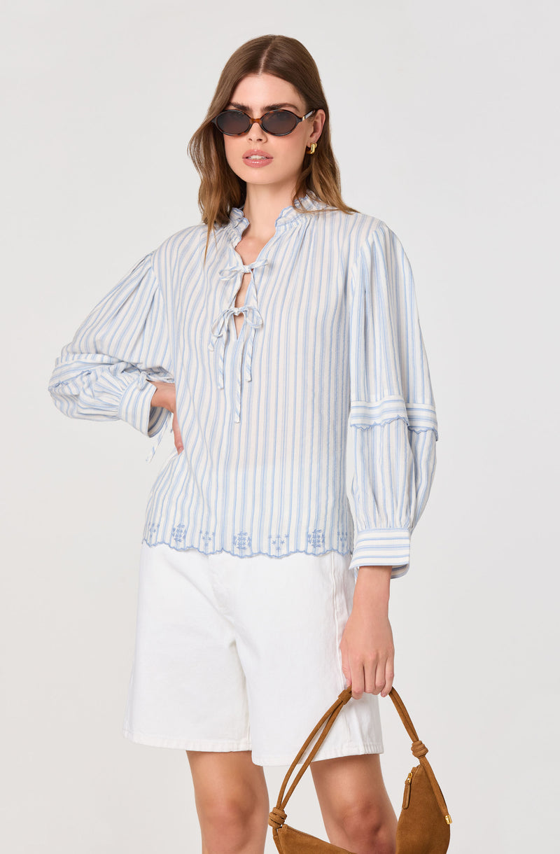 Woman in a light blue and white striped blouse with tie details and white shorts, holding a brown handbag.