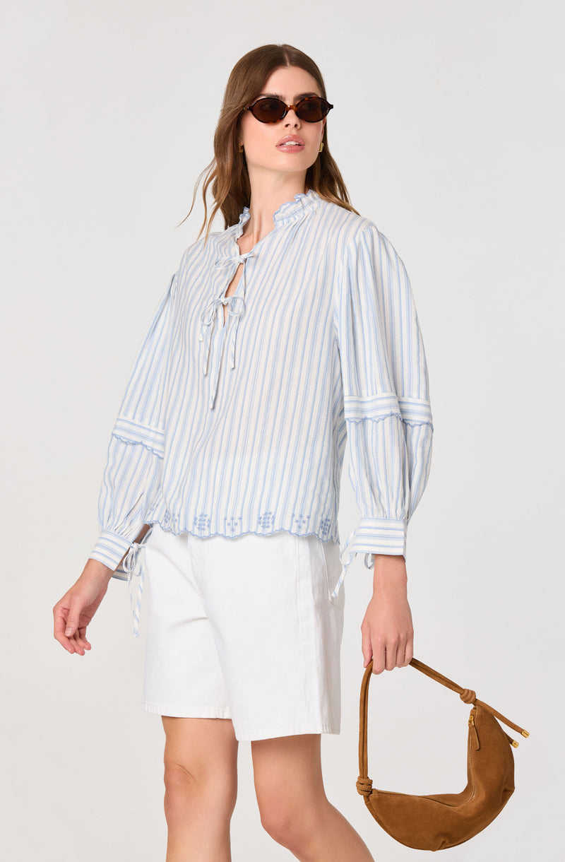Woman in a light blue and white striped blouse with a ruffled collar and tied neckline, paired with white shorts.