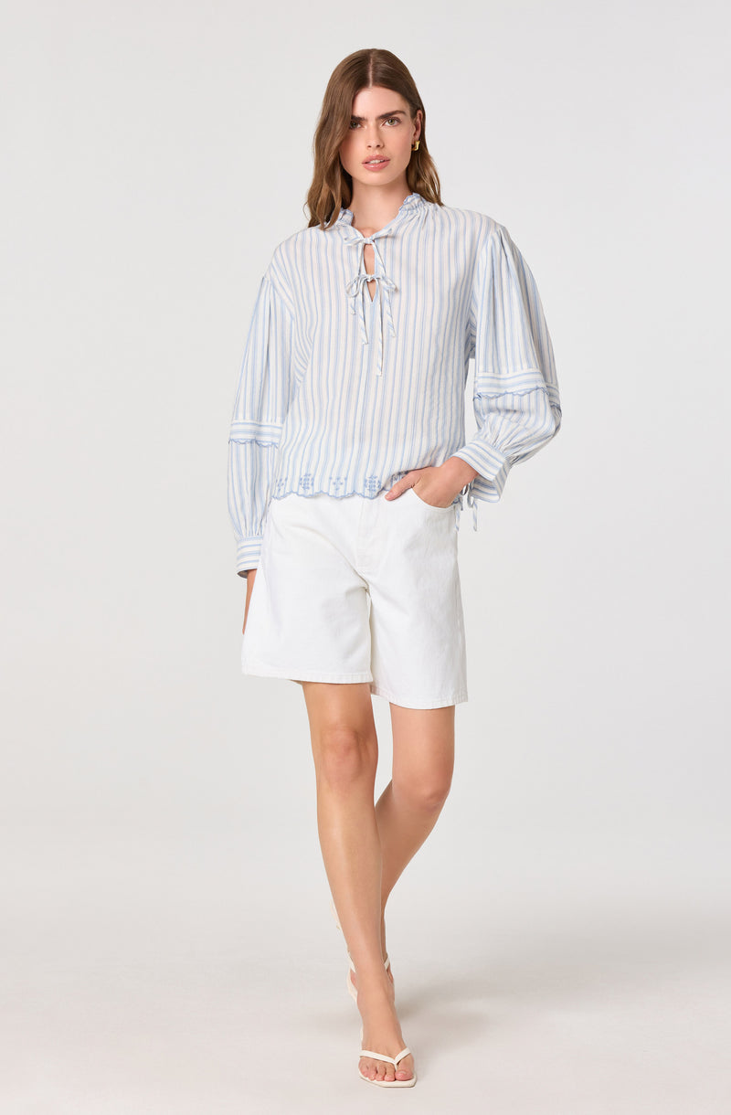 Woman in a striped light blue and white long-sleeve blouse with a tie detail, paired with white shorts.