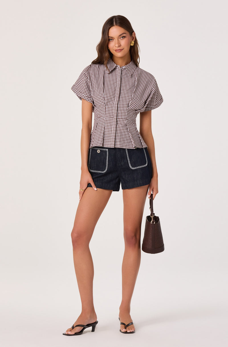 Girl wearing a checkered short-sleeve blouse, dark denim shorts with white stitching, black heeled sandals, and holding a brown handbag.
