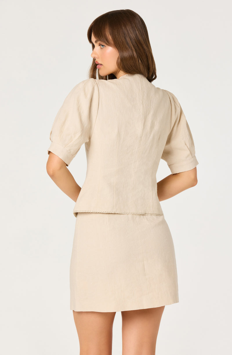 Woman in beige short-sleeve blouse with puffed sleeves and matching beige skirt.