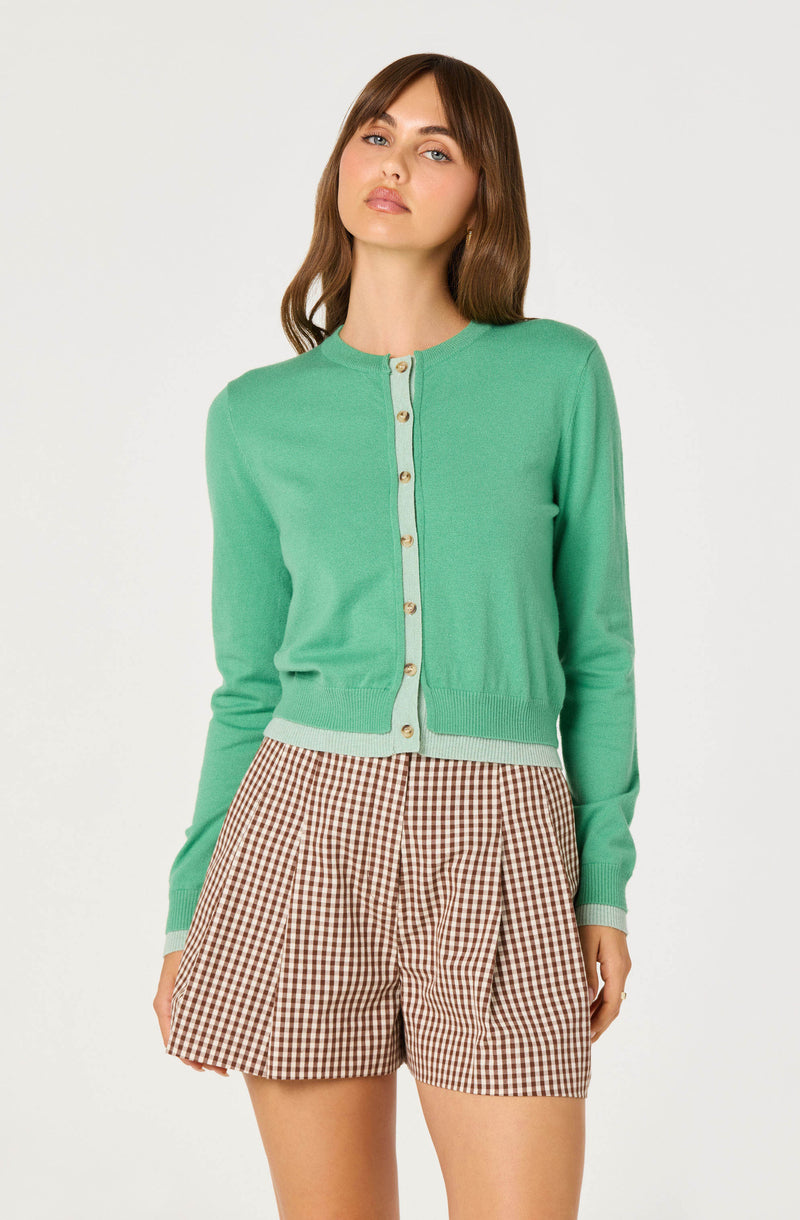 Woman wearing a light green cardigan with beige buttons and brown gingham shorts.
