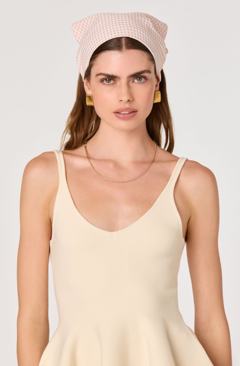 Woman wearing a pink and white checkered headscarf, gold square earrings, a delicate gold chain necklace, and a cream sleeveless dress.