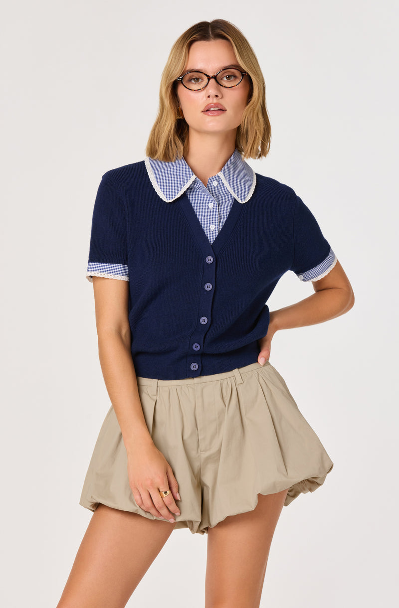 Woman wearing a navy blue short-sleeve cardigan with a blue and white checkered collar and sleeves, paired with beige puffed shorts.