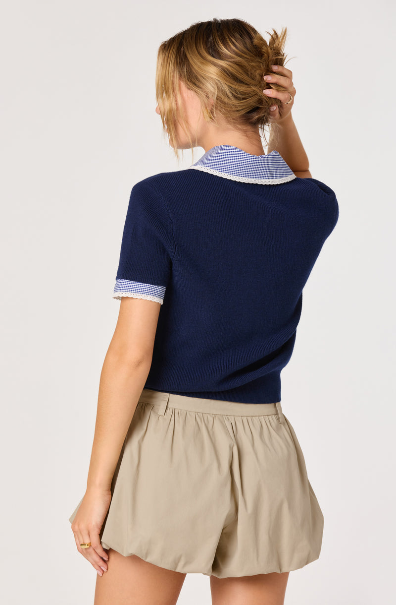 Woman wearing a navy blue sweater with light blue checkered collar and sleeve trim, beige shorts.