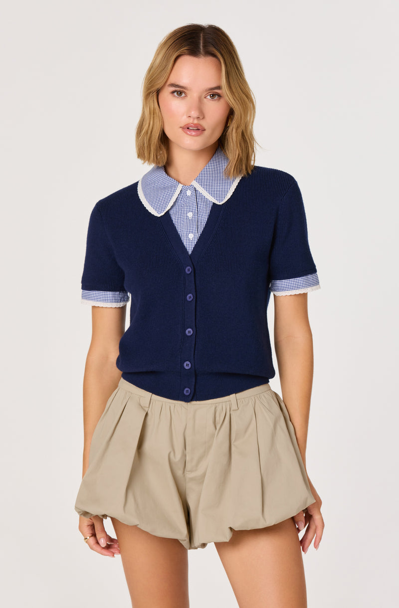 Woman wearing beige shorts and a navy blue cardigan over a blue and white checkered collared shirt.