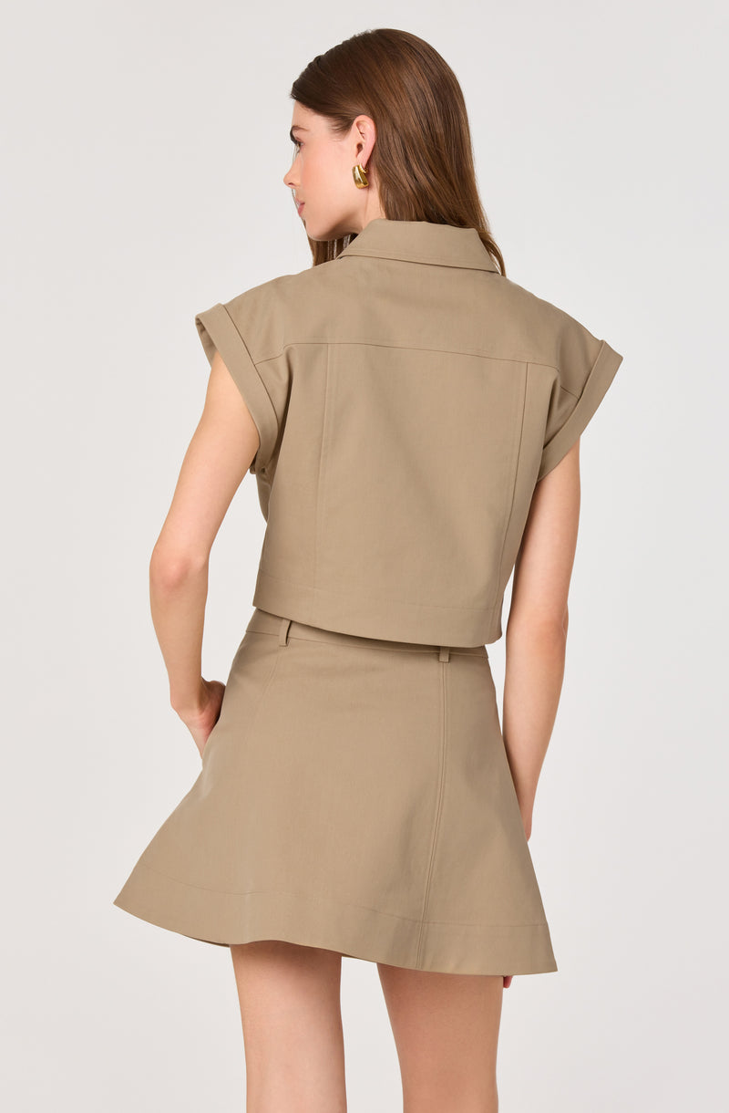 Woman wearing a beige sleeveless cropped jacket with rolled-up sleeves and a matching beige A-line skirt.