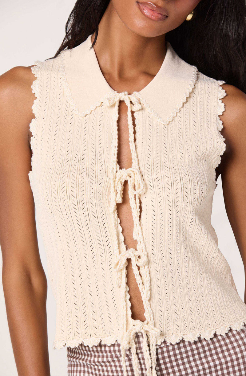 Cream sleeveless knit top with a Peter Pan collar, front tie closures, and ruffled edges.