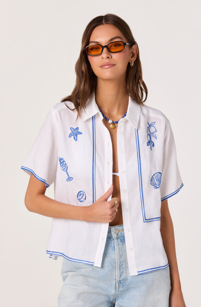 White short-sleeve button-up shirt with blue embroidered beach-themed designs, and blue piping.