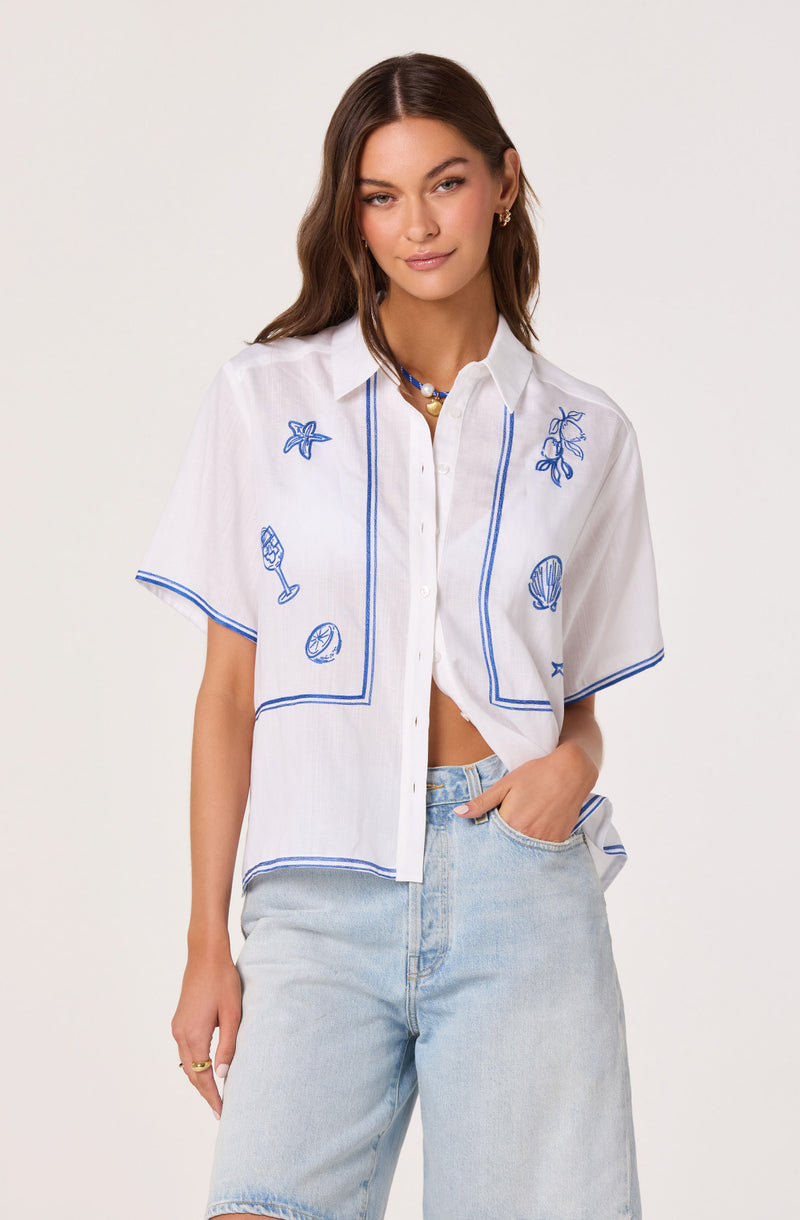 White short-sleeve shirt with blue embroidered beach and seashell designs, and light-wash denim shorts.