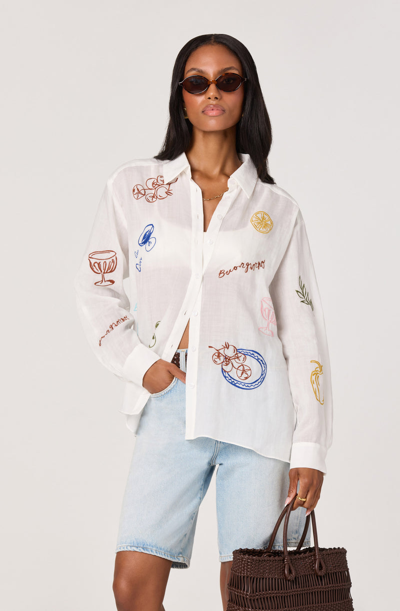 Woman wearing a white button-up shirt with colorful embroidered illustrations and text, paired with light blue denim shorts.