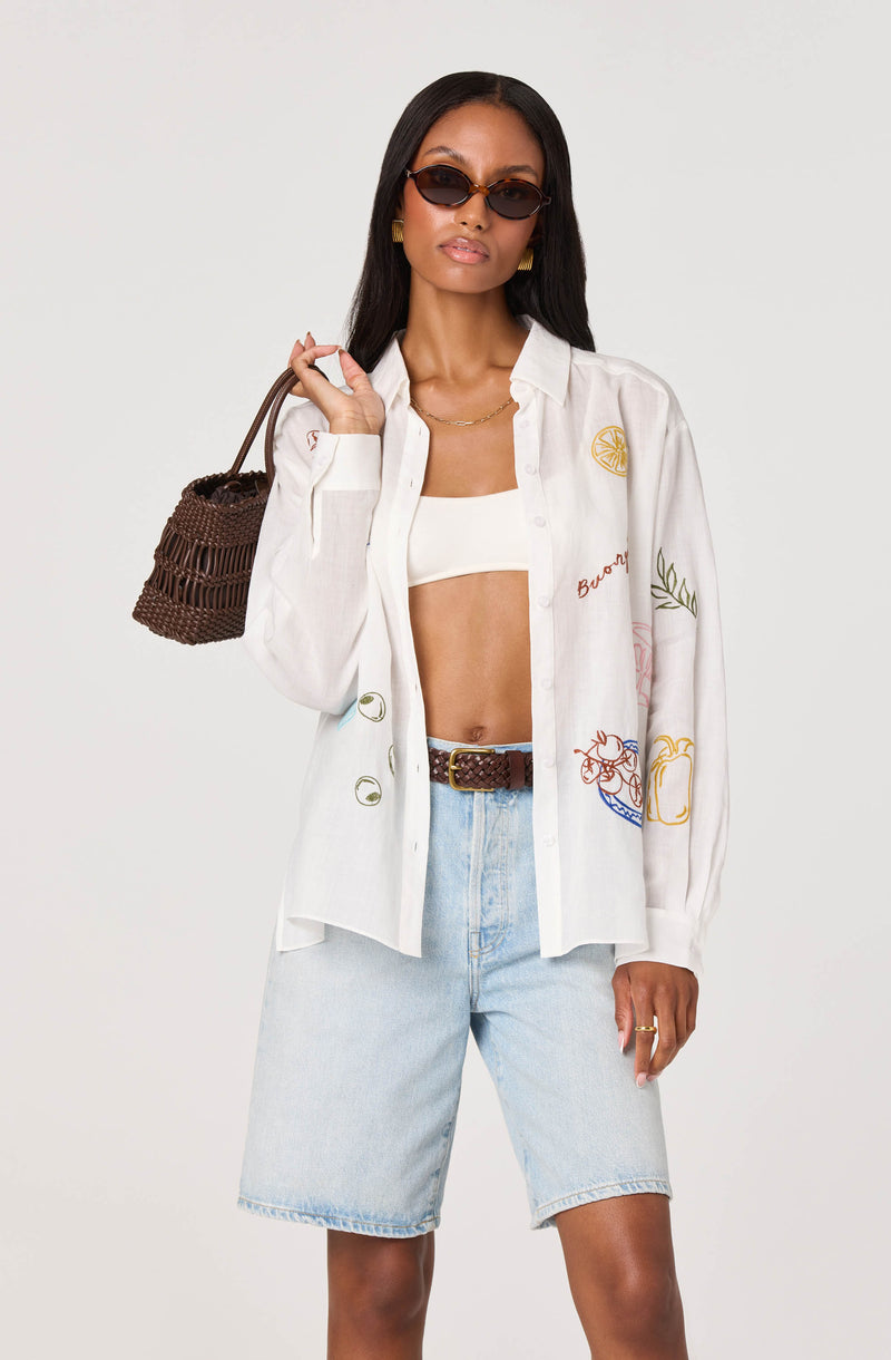 Woman wearing white embroidered button-up shirt, light blue denim shorts, brown belt, and sunglasses.