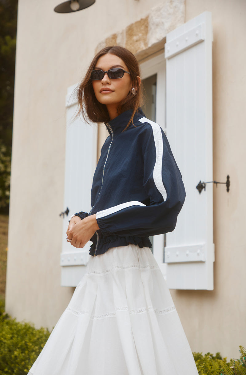 Woman wearing a navy blue windbreaker with white stripes and a long white skirt.