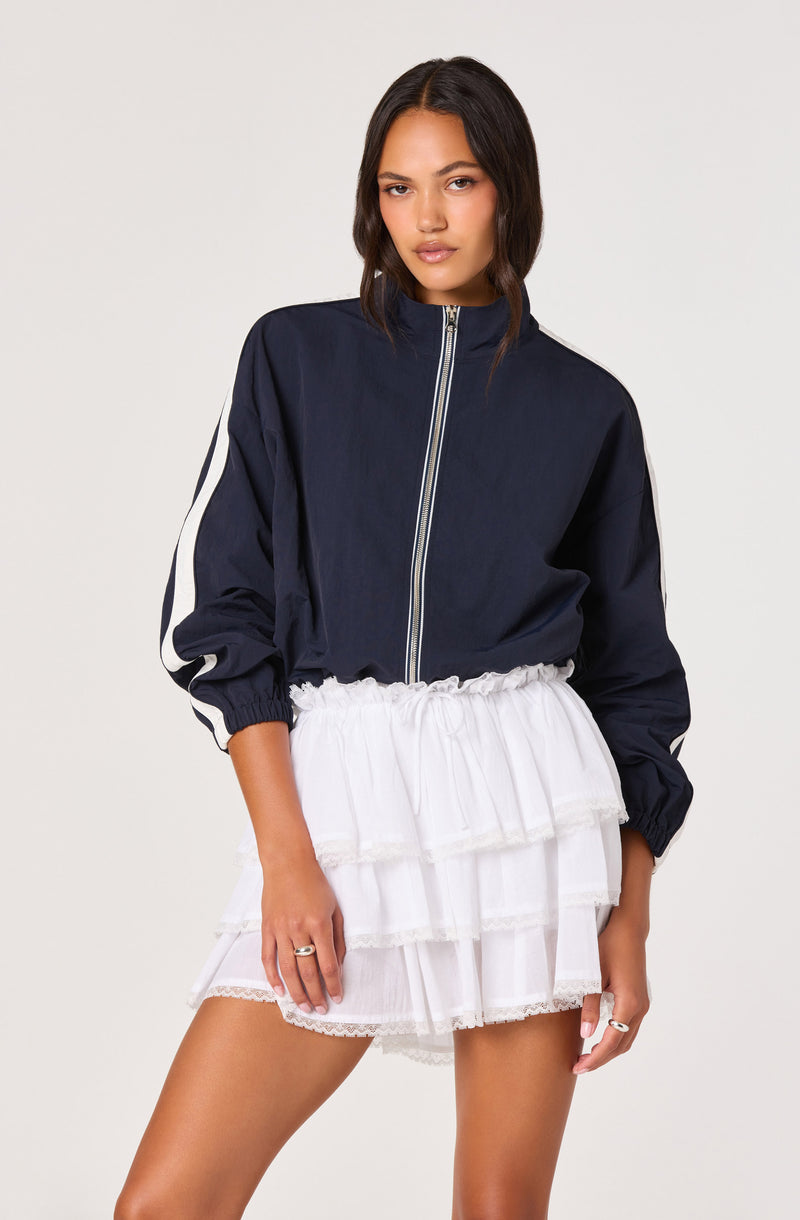 Model wearing a navy blue zip-up jacket with white stripe details on the sleeves and a white tiered ruffle skirt.