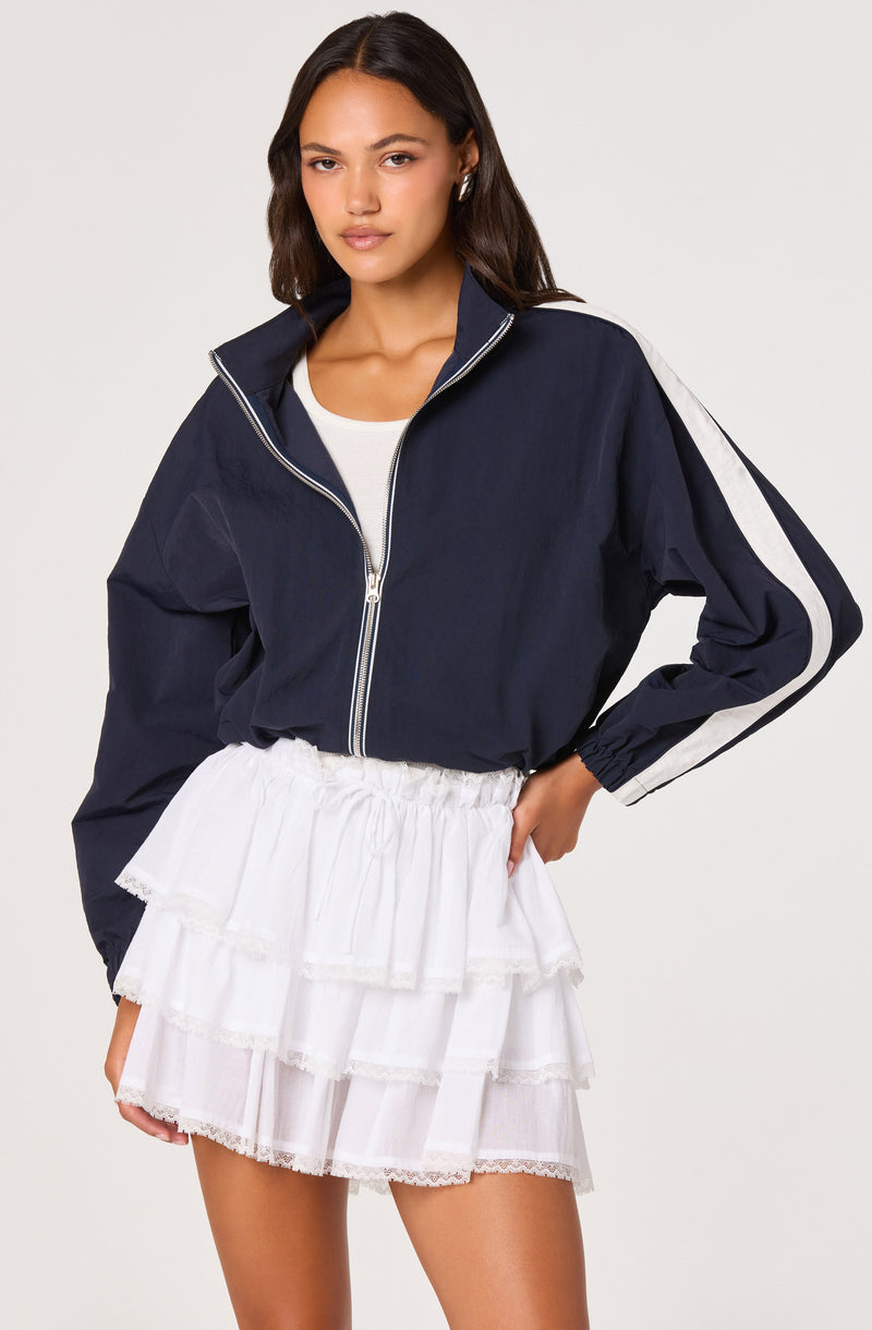 Woman wearing a navy zip-up jacket with white stripes on the sleeves and a white tiered skirt.
