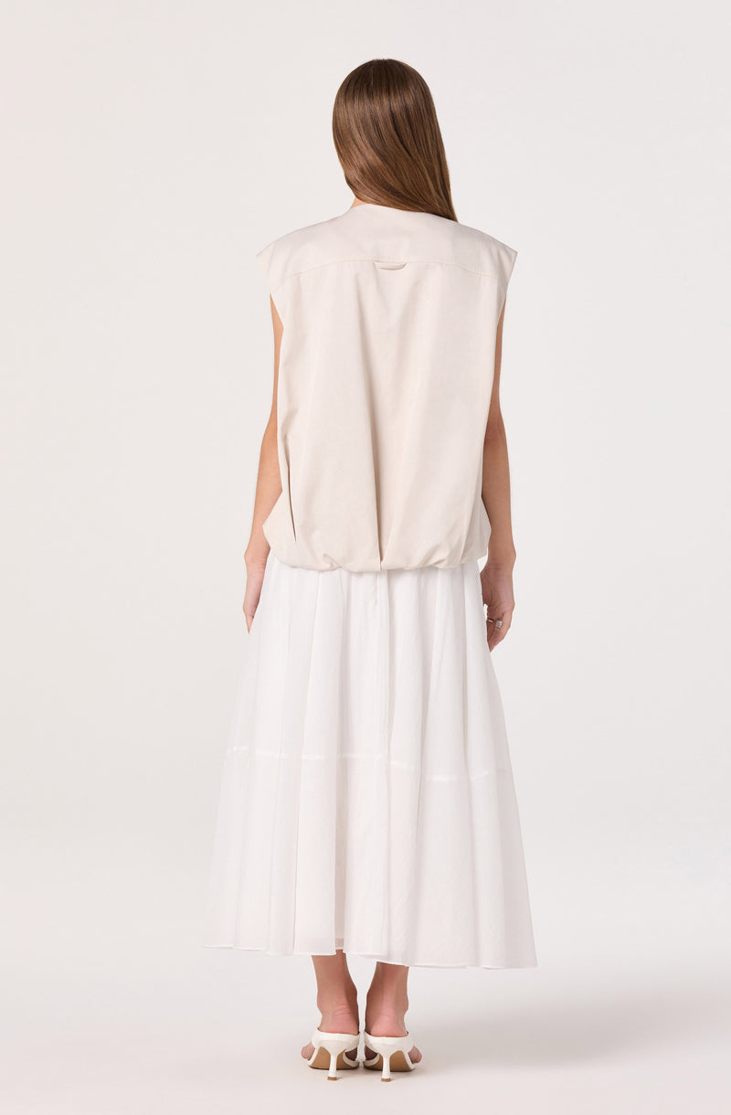 Woman in a sleeveless beige top and long white skirt, wearing white heels.