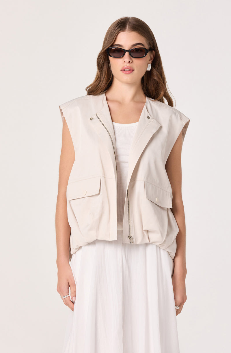 Woman wearing a sleeveless beige vest over a white top and white pleated skirt.