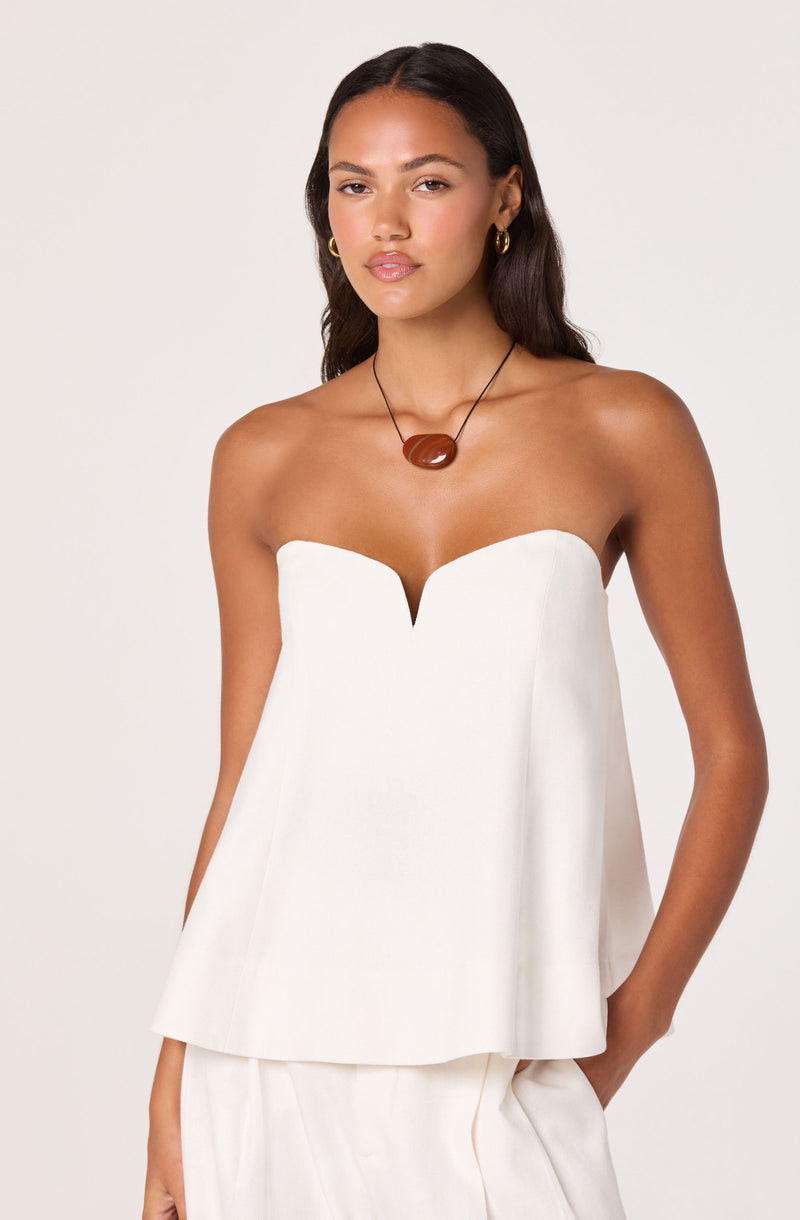 Woman in white strapless top with a deep V neckline, paired with matching pants, wearing gold earrings and a brown pendant necklace.