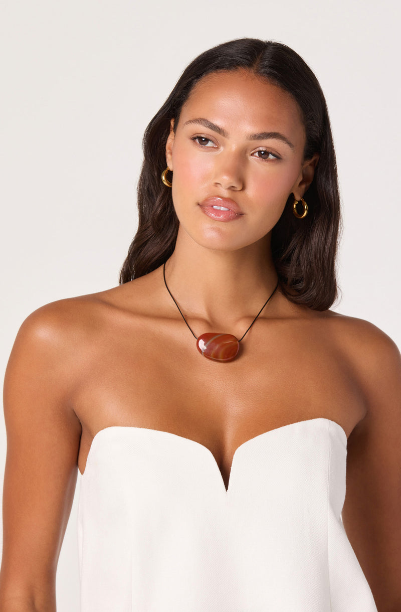 Woman wearing a strapless white top, gold hoop earrings, and a large red-brown stone necklace.
