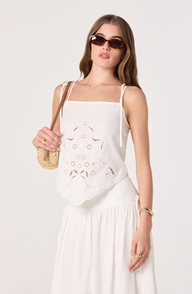 Young woman wearing a white, sleeveless embroidered top with scalloped hem and a white skirt.