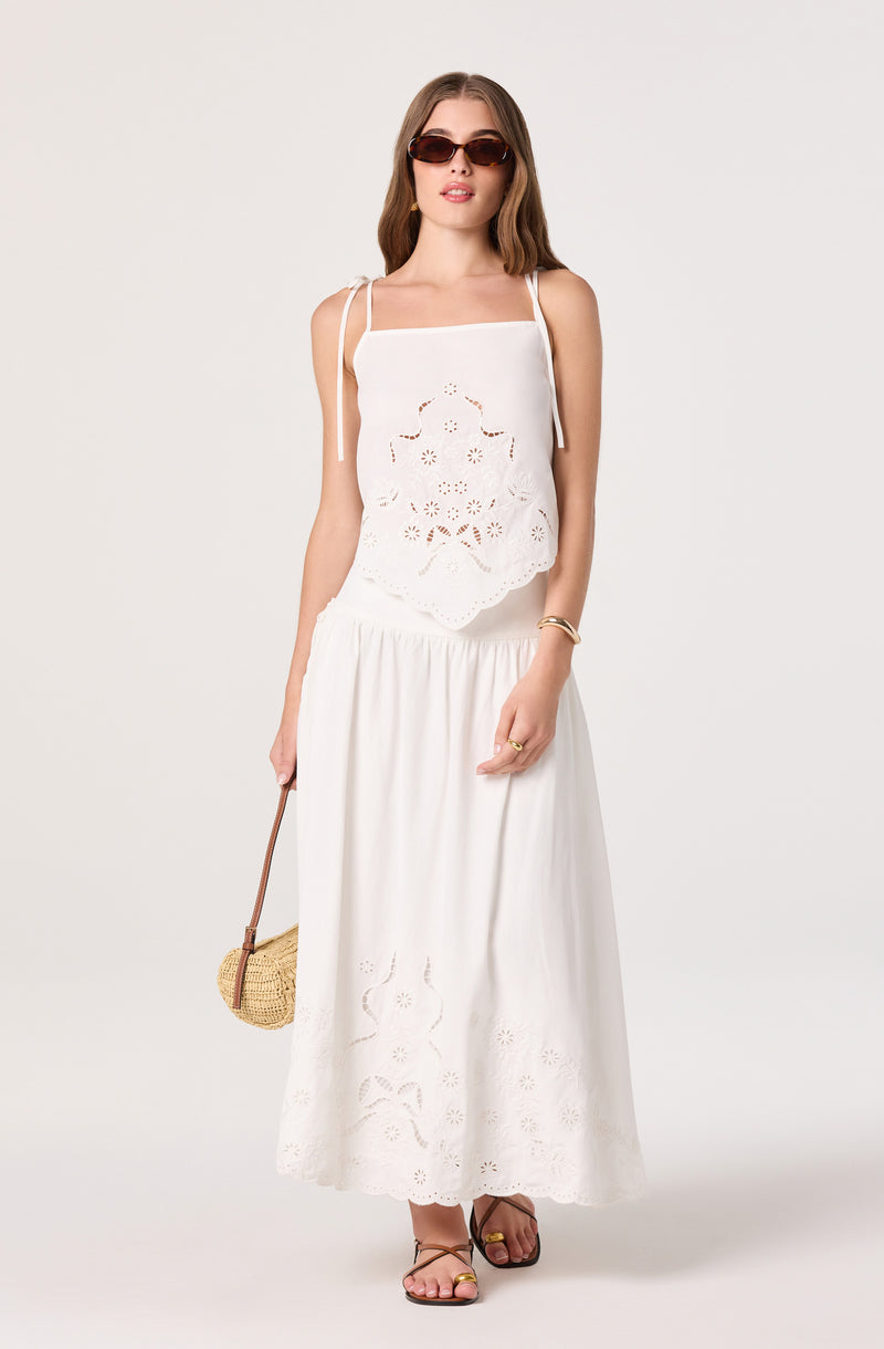 Woman wearing a white sleeveless top with tie straps and eyelet embroidery, and a long white skirt with eyelet details.