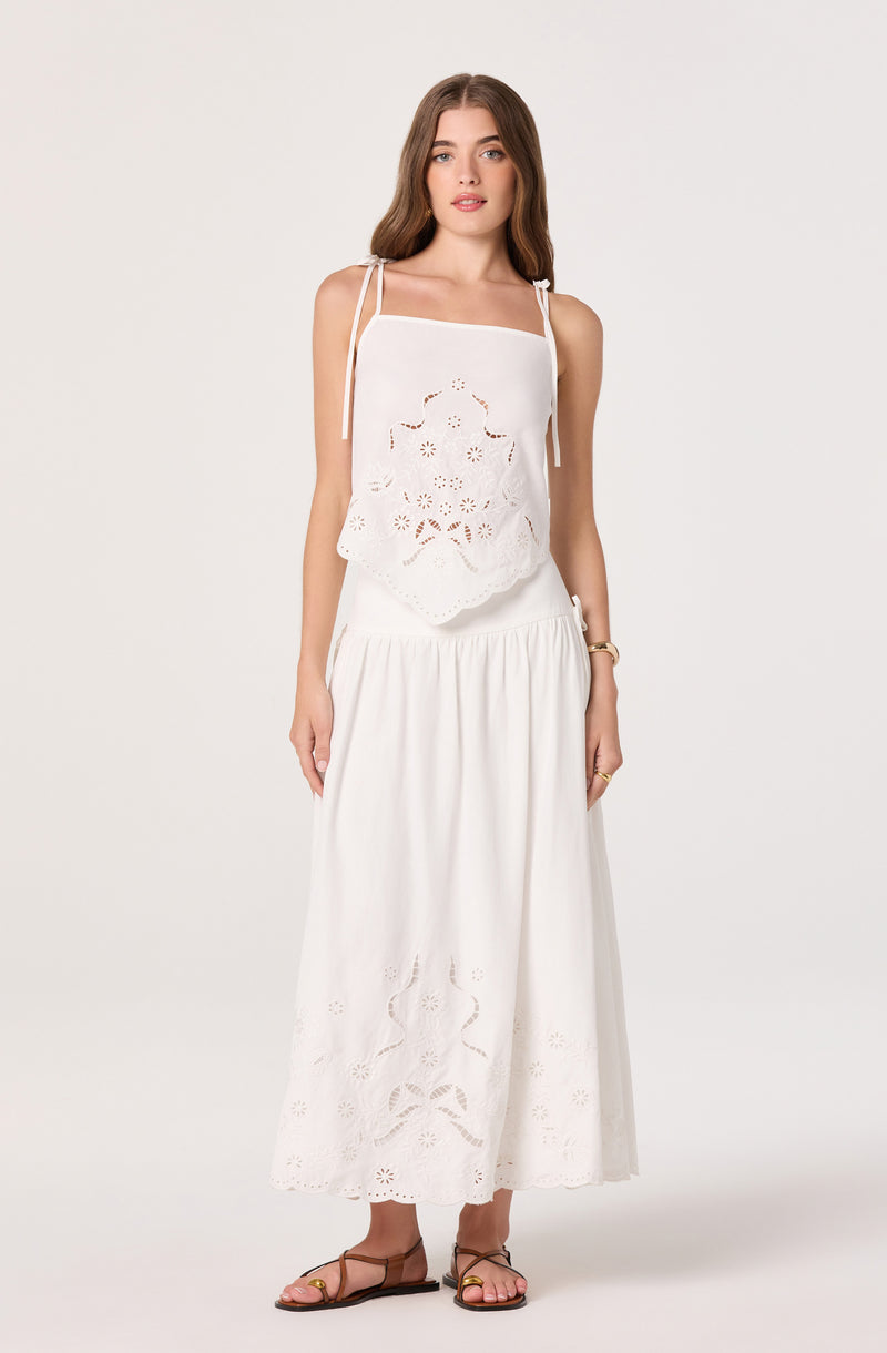 Woman wearing a white embroidered sleeveless top with shoulder tie straps and a long white skirt with eyelet details.