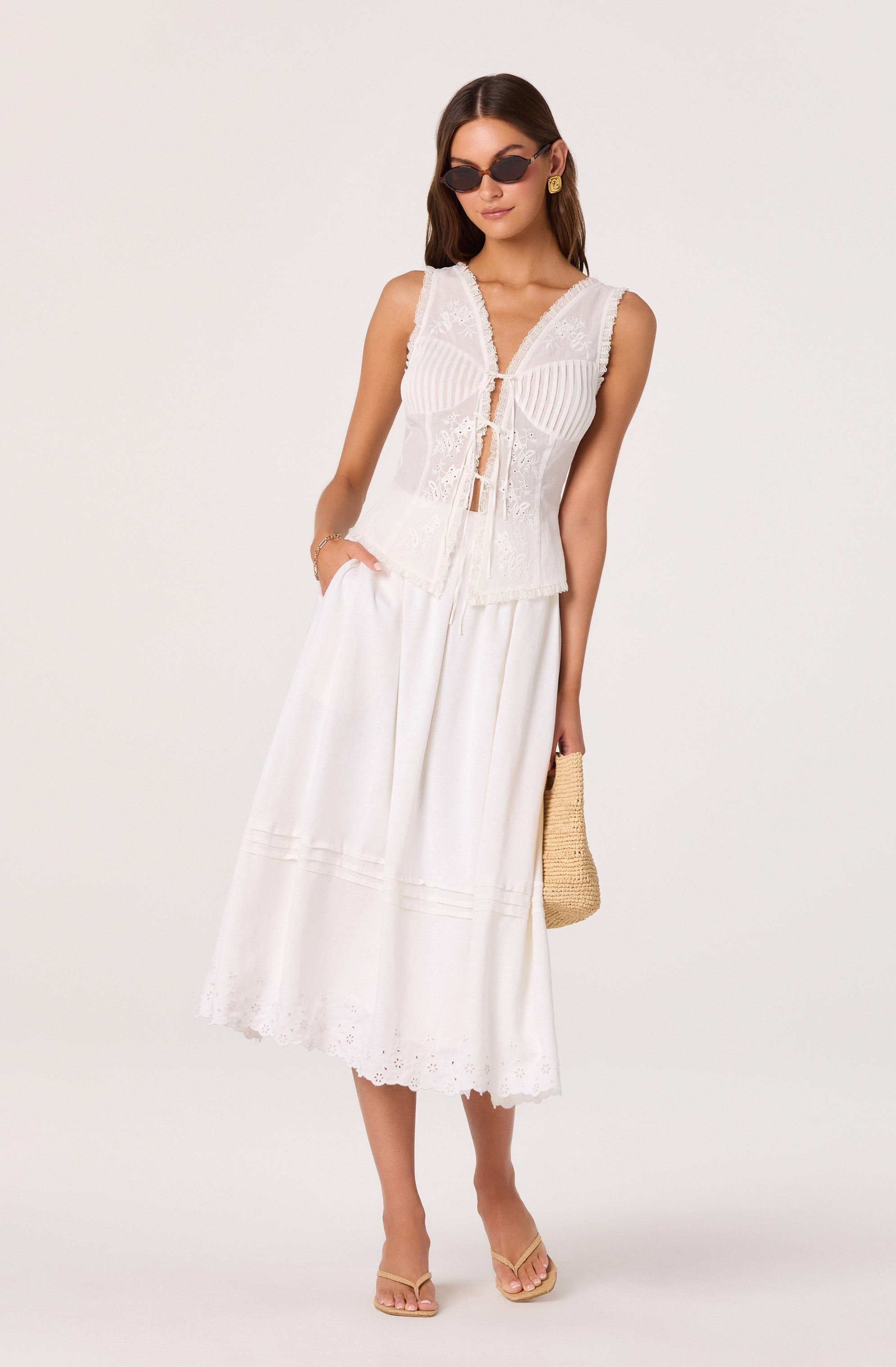 Woman wearing a sleeveless white embroidered lace top, and a long white skirts with eyelet embroidery. She's accessorized with sunglasses, gold earrings, a bracelet, and carries a straw handbag.