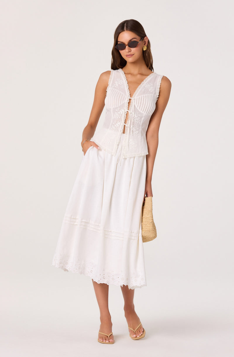 Woman wearing a sleeveless white embroidered lace top, and a long white skirts with eyelet embroidery. She's accessorized with sunglasses, gold earrings, a bracelet, and carries a straw handbag.