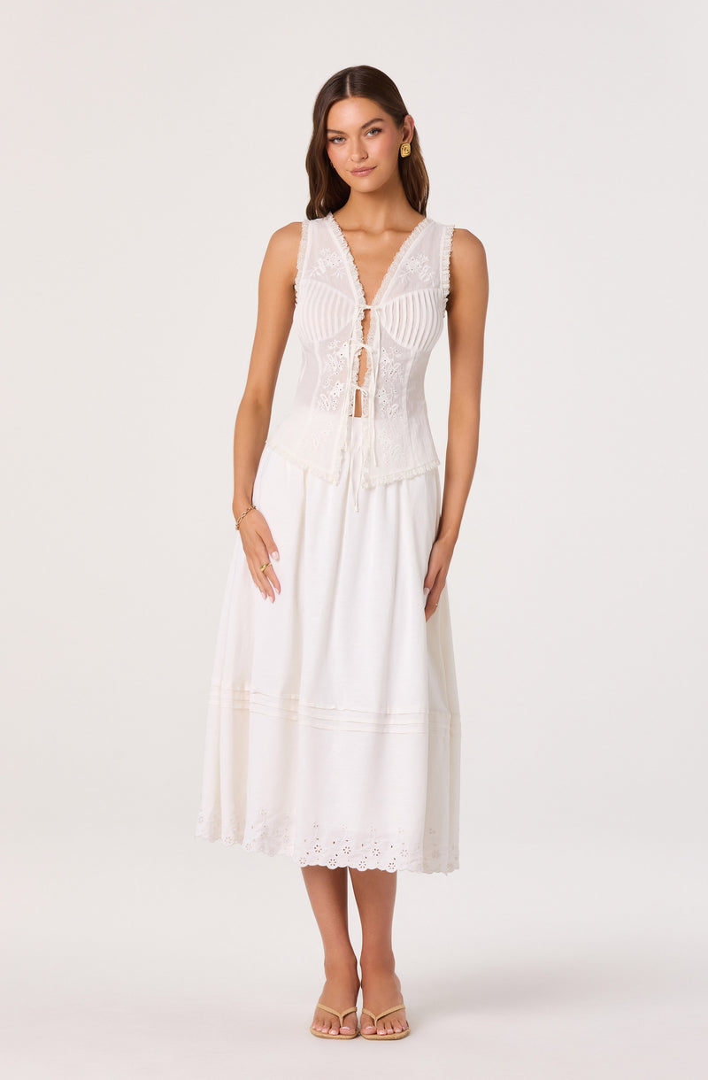 Woman wearing a sleeveless white top with lace details and a tie-front, paired with a white, ankle-length skirt.