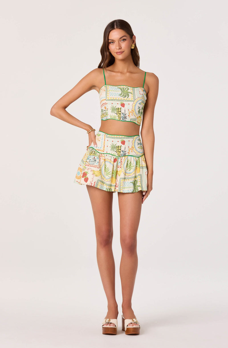 Young woman in a colorful tropical print crop top with thin green straps and matching ruffled skirt, beige platform sandals