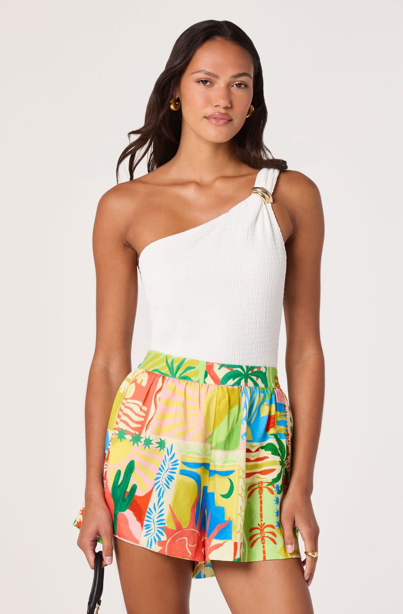 Woman wearing a one-shoulder white top with gold embellishment and colorful tropical print shorts.
